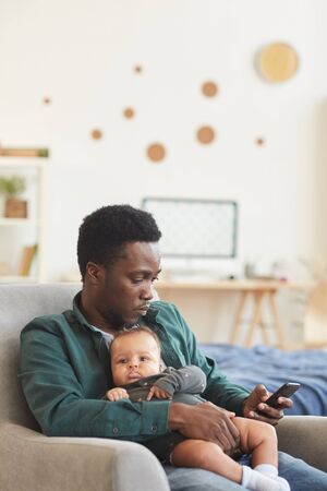 Vertical portrait of young African-American father holding cute mixed-race baby while sitting in comfortable armchair and using smartphone, copy spaceの写真素材