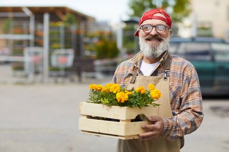 Waist up portrait of cheerful senior gardener holding flowers and looking at camera while standing by tree market outdoors, copy spaceの写真素材