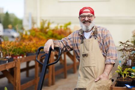 Waist up portrait of cheerful senior gardener looking at camera while leaning on shovel enjoying...