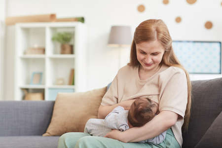 Portrait of happy mature mother breastfeeding baby while sitting on couch in home interior, copy...