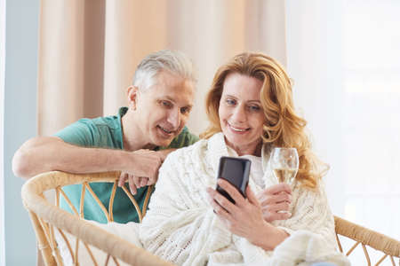 Warm-toned portrait of elegant mature couple looking at smartphone screen while relaxing at home,...