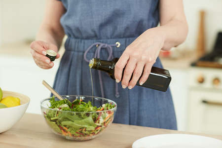 Close up of unrecognizable elegant woman adding olive oil to healthy salad in glass bowl while enjoying cooking in home kitchen, copy spaceの写真素材