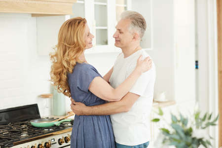 Waist up portrait of smiling mature couple embracing and looking at each other while standing in...