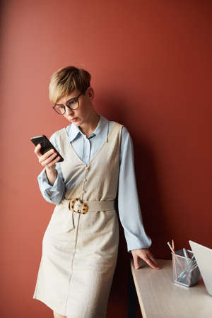 Vertical portrait of contemporary short-hired businesswoman using smartphone while standing against...
