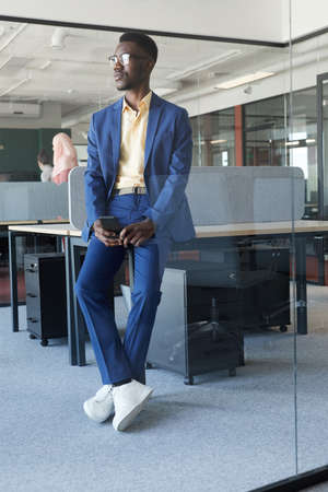 Vertical full length portrait of successful African-American businessman looking away while leaning...