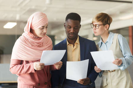 Waist up portrait of young multi-ethnic business team holding documents while discussing work project in officeの写真素材