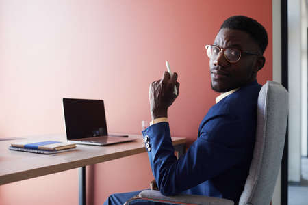 Portrait of elegant African-American man glancing at camera while using laptop at desk while working in contemporary office interior against red wall, copy spaceの写真素材