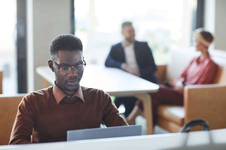 Portrait of young African-American man dressed in business casual using laptop in office or...