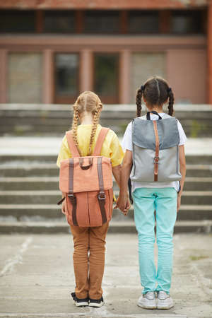 Full length back view portrait of two cute schoolgirls with backpacks standing outdoors against school building and holding handsの写真素材
