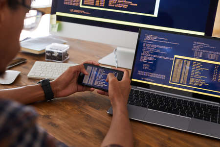 Close up of male hands holding smartphone with code on screen while working at desk in office, IT developer concept, copy spaceの写真素材
