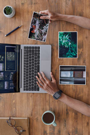 Minimal background composition of male hands holding printed photographs and using laptop on textured wooden desk, photographers office, copy spaceの写真素材