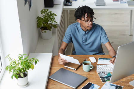 High angle portrait of creative African-American man reviewing photographs while working on editing...