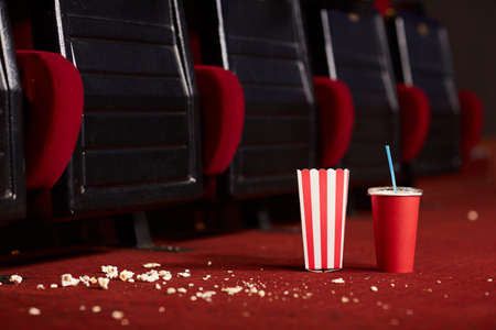 Close up background image of soda cup and popcorn on messy red floor in empty cinema theater, copy...