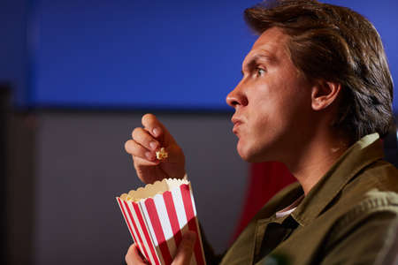 Side view portrait of young man eating popcorn while watching movie in cinema theater alone, copy...
