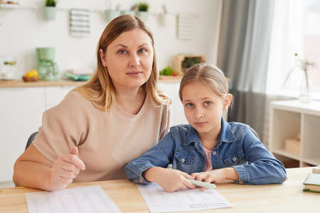 Warm-toned portrait of adult mother helping daughter doing homework while studying at home and looking at cameraの写真素材