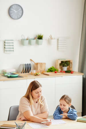 Vertical warm-toned portrait of caring mother helping daughter doing homework and studying at home in cozy interior, copy spaceの写真素材