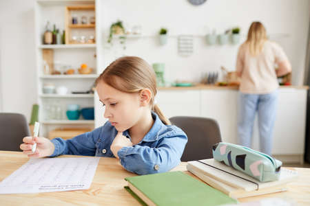 Warm-toned portrait of cute little girl doing homework while sitting at desk in cozy interior with mother in background, copy spaceの写真素材