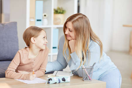 Portrait of loving adult mother looking at cute little girl doing homework while studying at home in...