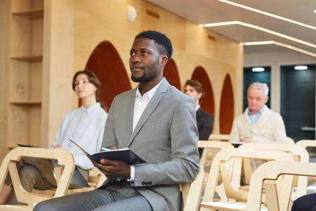 Portrait of African-American businessman taking notes while sitting in audience of contemporary...