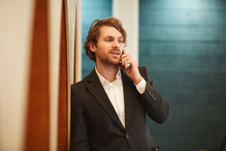 Waist up portrait of handsome bearded businessman speaking by smartphone and looking away while leaning on wall in minimal office interior, copy spaceの写真素材