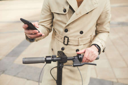 Cropped portrait of unrecognizable young man wearing trenchcoat holding smartphone while riding electric scooter in urban city setting, copy spaceの写真素材