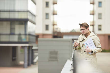 Minimal waist up portrait of modern young businessman wearing trenchcoat and holding smartphone...