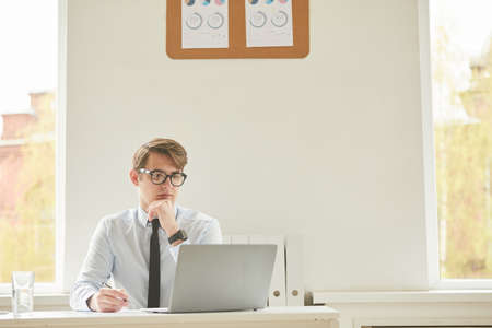 Minimal portrait of young businessman wearing glasses and using laptop while working at desk in...