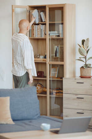 Vertical back view at bald person putting book on shelf on wooden bookcase in modern home interior, alopecia and cancer awareness, copy spaceの写真素材