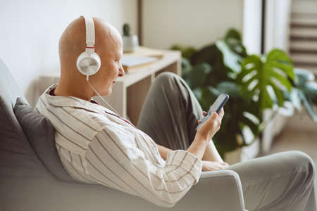 Minimal back view portrait of bald adult woman wearing headphones while listening to music via smartphone in modern home interior, alopecia and cancer awareness, copy spaceの写真素材