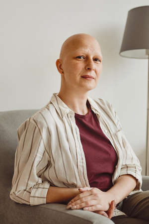 Vertical warm-toned portrait of confident bald woman looking at camera while sitting on couch in minimal home interior, alopecia and cancer awarenessの写真素材