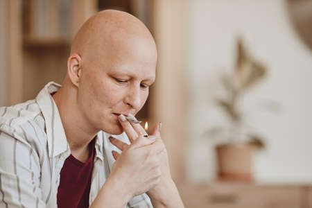 Warm-toned portrait of bald adult woman smoking marijuana joint for medicinal purposes in cancer...