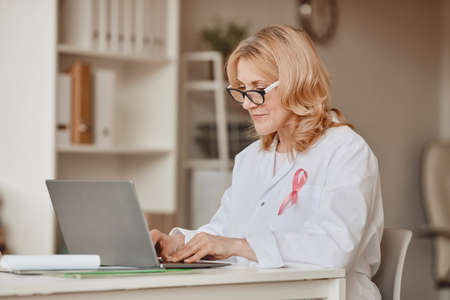 Warm-toned portrait of mature female doctor with pink ribbon pinned on white lab coat while working at laptop in modern office, breast cancer awareness symbol, copy spaceの写真素材
