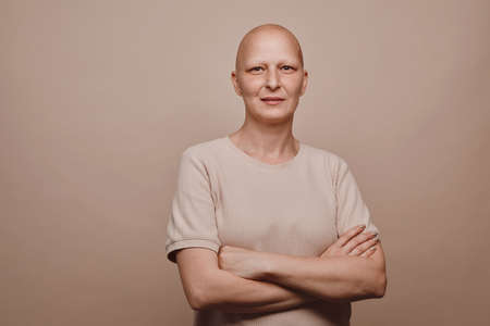 Warm-toned waist up portrait of confident bald woman looking at camera while posing against minimal beige background in studio, alopecia and cancer awareness, copy spaceの写真素材