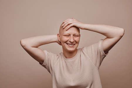 Warm-toned waist up portrait of carefree bald woman touching shaved head and smiling while posing against minimal beige background in studio, alopecia and cancer awareness, copy spaceの写真素材
