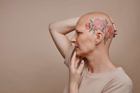 Minimal side view portrait of bald woman with head tattoo posing against beige background in studio, alopecia and cancer awareness, copy spaceの写真素材