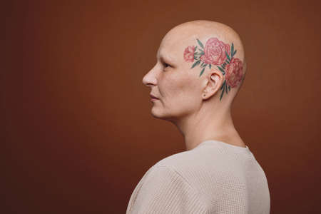Minimal side view portrait of confident bald woman with head tattoo posing against brown background...