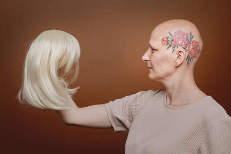 Side view portrait of confident bald woman holding wig of blonde hair against brown background in studio, alopecia and cancer awareness, copy spaceの写真素材