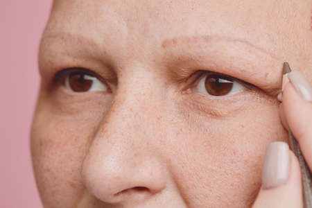 Extreme close up of freckled bald woman drawing eyebrows and doing makeup against pink background in studio, alopecia and cancer awareness, copy spaceの写真素材