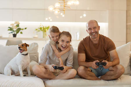 Cute warm-toned portrait of happy modern family playing video games together while sitting on couch...
