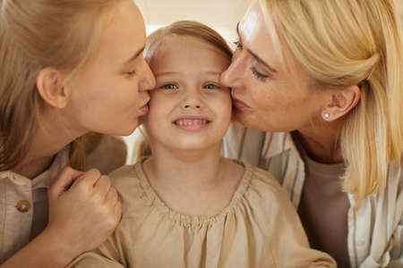 Warm-toned close up portrait of mother and sister kissing cute little girl on both cheeks, family bonds and generations conceptの写真素材