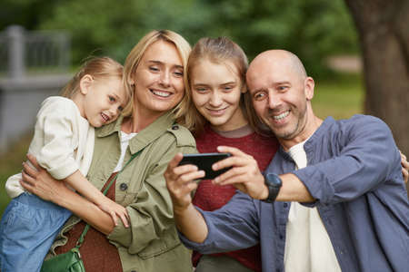Waist up portrait of modern happy family with two daughters taking selfie outdoors via smartphone...