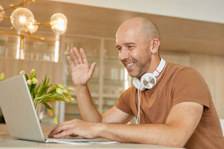 Warm-toned portrait of bald mature man smiling and waving at camera while speaking by video chat via...