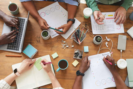 Top view close up of multi-ethnic group of people working together at cluttered wooden table with coffee cups, mugs and stationary items, teamworking or studying concept, copy spaceの写真素材