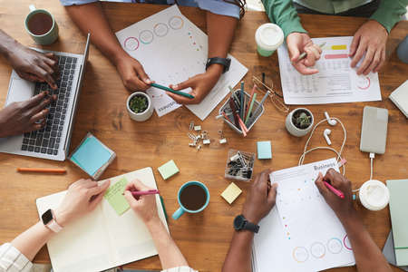 Top view close up of multi-ethnic group of people working together at cluttered wooden table with coffee cups, mugs and stationary items, teamworking or studying concept, copy space