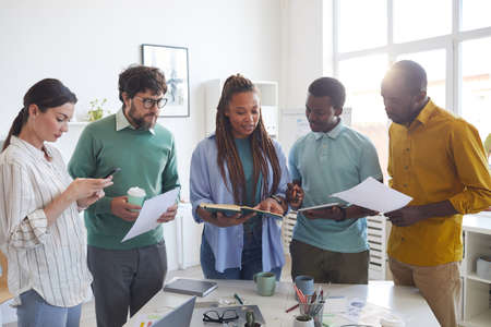 Portrait of contemporary multi-ethnic business team standing around table in office and listening to...