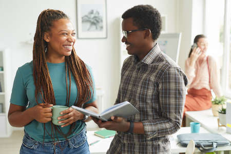 Waist up portrait of contemporary African-American woman talking to colleague and smiling cheerfully...