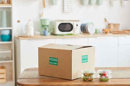 Background image of cardboard box with food standing on wooden table in white kitchen interior, food...