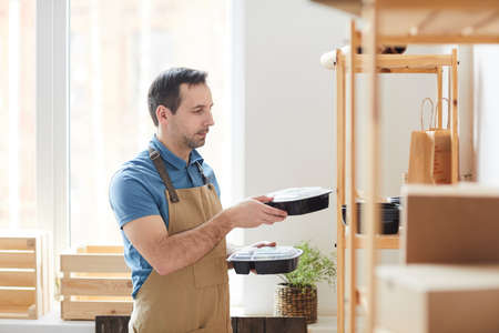 Side view portrait of mature man wearing apron stacking plastic packaging on shelves while working in food delivery service, copy spaceの写真素材