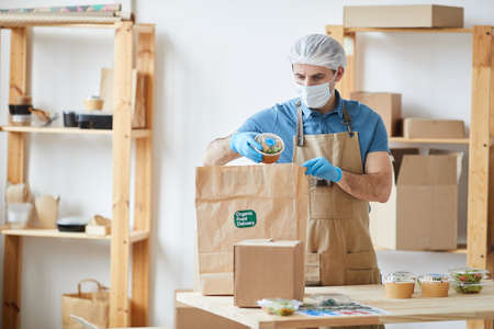 Waist up portrait of mature male worker wearing protective clothes while safely packaging orders at wooden table in food delivery service, copy spaceの写真素材