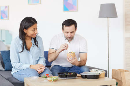 Portrait of smiling man and woman opening food delivery containers while enjoying takeout lunch in...